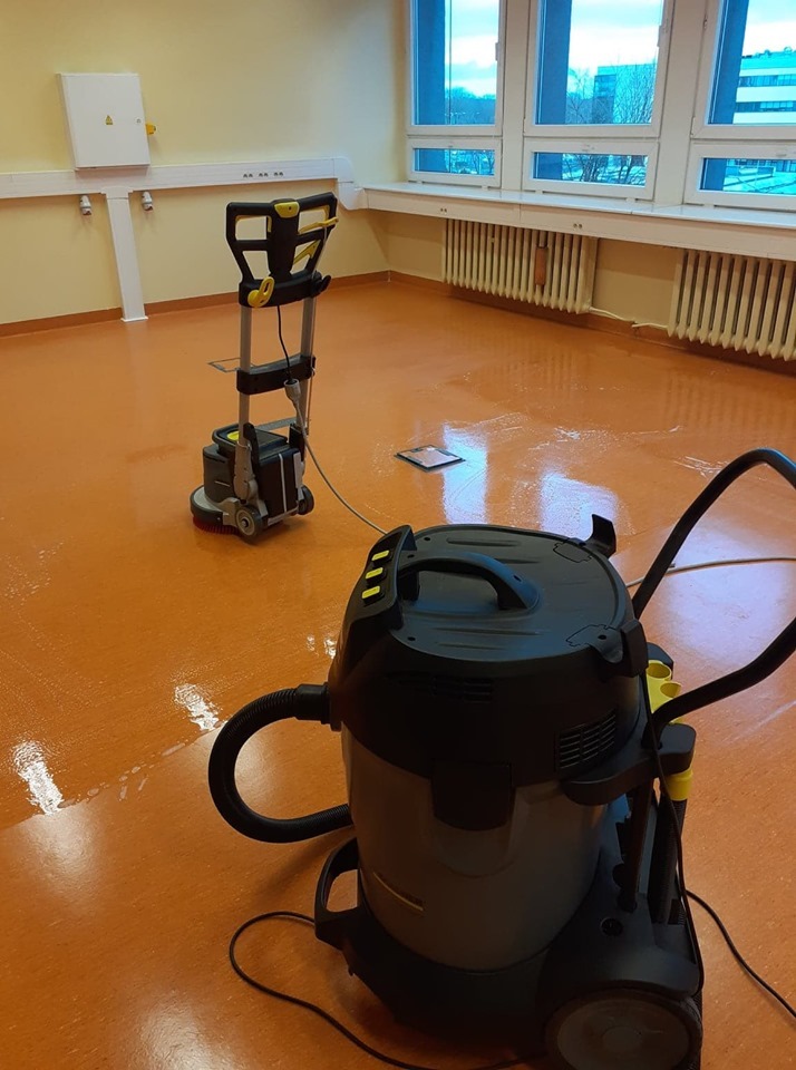 Pomarańczowa podłoga w biurze po umyciu, widoczny odkurzacz przemysłowy i automat do mycia podłóg, w tle okno z widokiem na budynek.