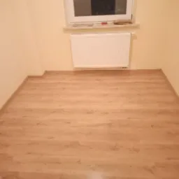 Jasne, drewnopodobne panele podłogowe w pokoju z beżowymi ścianami, biały grzejnik pod oknem.