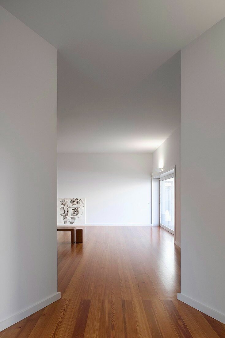 Wnętrze minimalistycznego mieszkania z drewnianą podłogą i białymi ścianami, widok z korytarza na salon z abstrakcyjnym obrazem i przesuwnymi drzwiami balkonowymi.