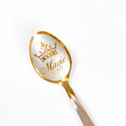 Złota łyżeczka z grawerem korony i napisem 'Magie' na białym tle.
