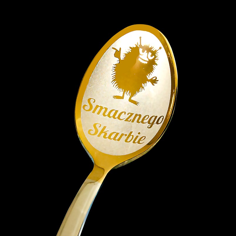 Złota łyżeczka z wygrawerowanym napisem 'Smacznego Skarbie' i rysunkiem zabawnego potworka na czarnym tle.