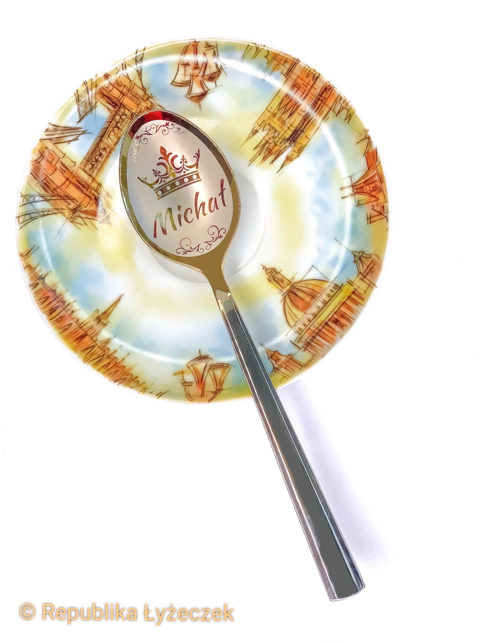 Łyżeczka z grawerowanym imieniem 'Michał' i koroną, leżąca na talerzyku z motywem architektonicznym w odcieniach brązu i błękitu.