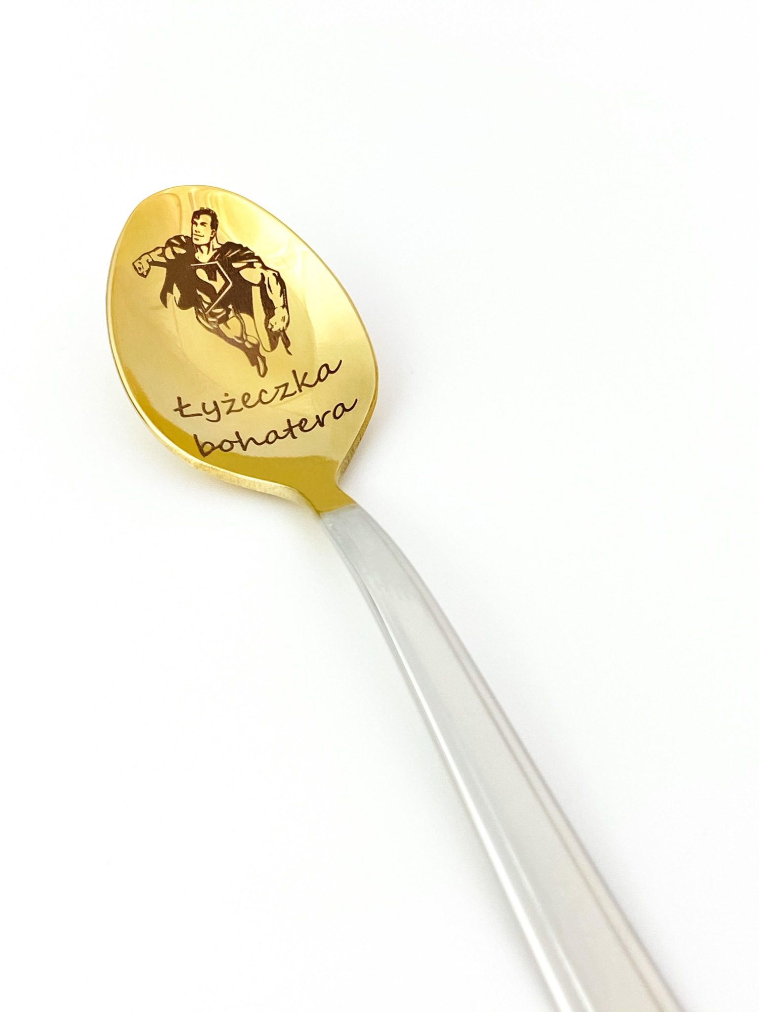 Złota łyżeczka z wygrawerowaną postacią Supermana i napisem 'łyżeczka bohatera', na białym tle.