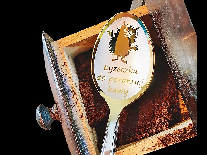 Łyżeczka z napisem 'Łyżeczka do porannej kawy' i rysunkiem zabawnego stworka, umieszczona w drewnianej szufladce wypełnionej zmieloną kawą.