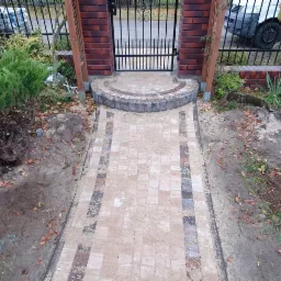 Nowo ułożona ścieżka z kostki brukowej prowadząca do furtki, z widocznymi śladami prac ziemnych i przygotowanymi roślinami do nasadzeń.