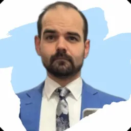 Portret mężczyzny w niebieskiej marynarce i krawacie w kwiaty, z lekkim zarostem, na rozmytym tle z elementami niebieskiego i białego.