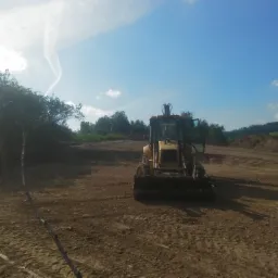 Żółta ładowarka na placu budowy z otwartymi drzwiami kabiny, świeżo wyrównany teren, w tle drzewa i błękitne niebo z smugami kondensacyjnymi.