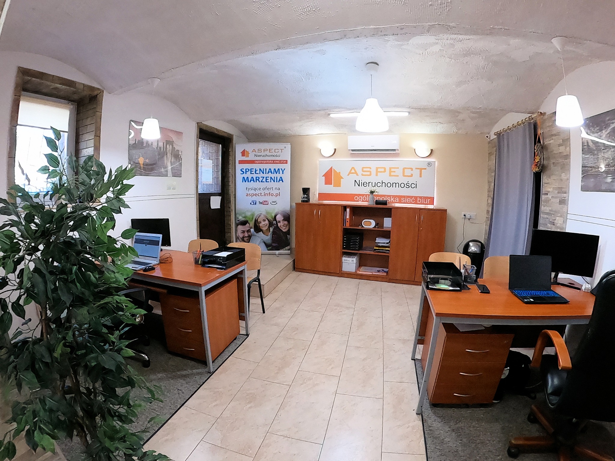 Wnętrze biura agencji nieruchomości z biurkami, komputerami, szafką i rośliną doniczkową; w tle widoczny baner reklamowy z logo i hasłem 'Spełniamy marzenia'.