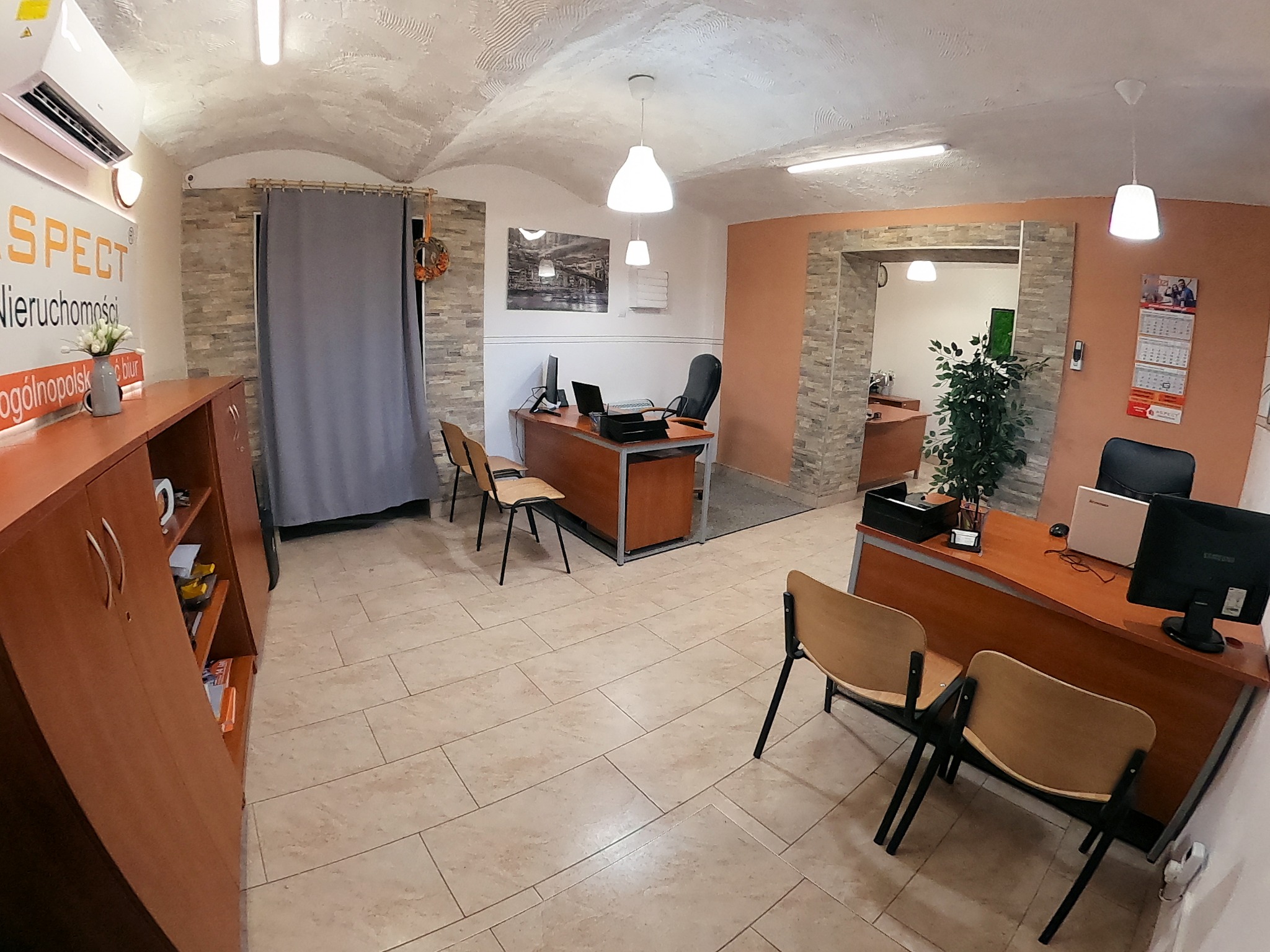 Wnętrze biura agencji nieruchomości w Piekarach Śląskich z drewnianymi meblami, biurkami z komputerami i krzesłami dla klientów, oświetlone wiszącymi lampami.