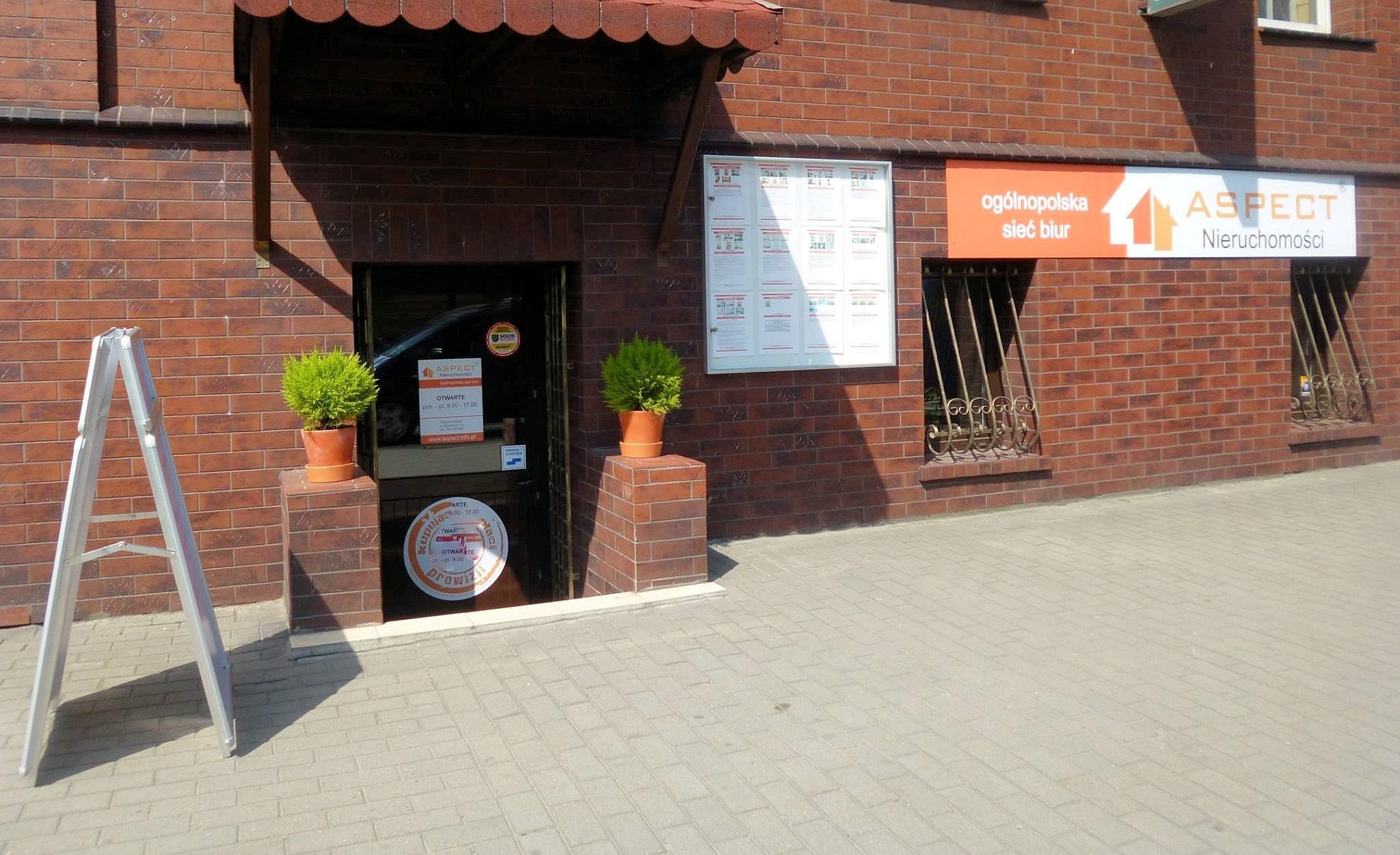 Wejście do biura nieruchomości Aspect z szyldem, tablicą informacyjną i stojakiem reklamowym na chodniku przed budynkiem z cegły.