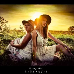 Dwie kobiety w białych, koronkowych sukienkach i kapeluszach siedzą plecami do siebie na łące o zachodzie słońca.