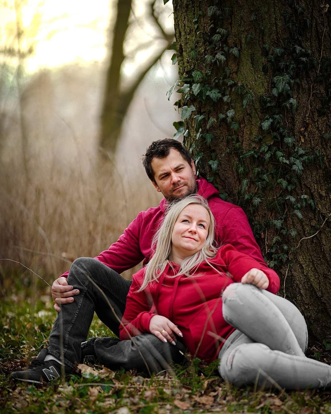 Para w czerwonych bluzach z kapturem siedzi oparta o drzewo porośnięte bluszczem w lesie, z delikatnym rozmyciem tła.