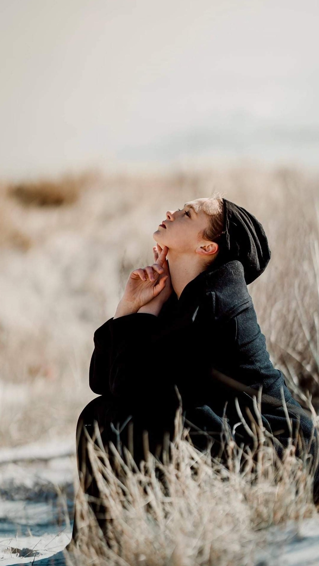 Młoda kobieta w ciemnym płaszczu i chuście na głowie siedzi w trawie, spoglądając w górę z rękami złożonymi pod brodą, w naturalnym oświetleniu.