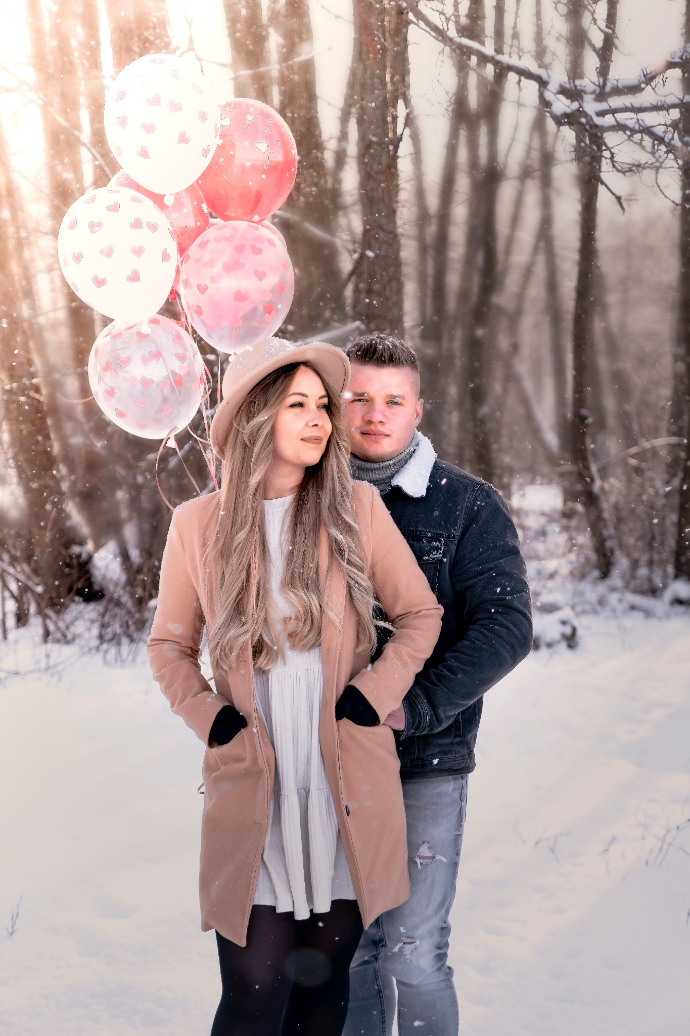 Para w zimowej scenerii, ona w beżowym płaszczu i kapeluszu trzyma balony z serduszkami, on w dżinsowej kurtce, delikatne promienie słońca przebijają przez drzewa.