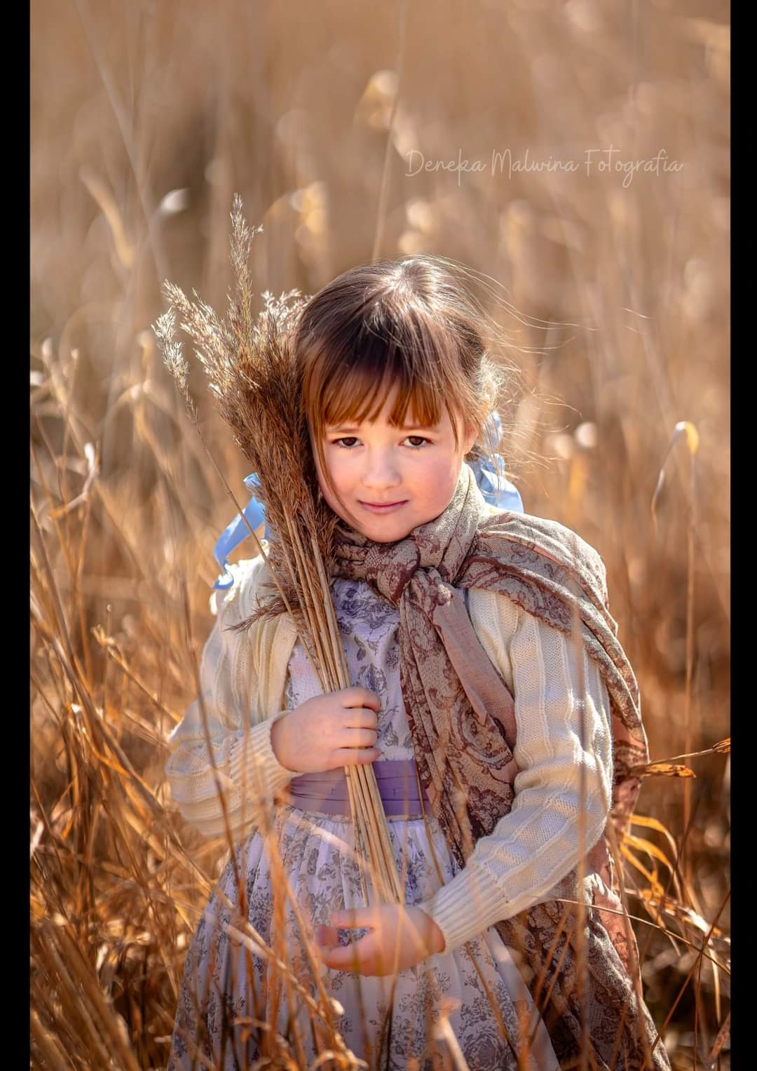 Portret dziewczynki z brązowymi włosami i grzywką, ubranej w kwiecistą sukienkę, kremowy sweterek i wzorzysty szal, trzymającej bukiet ususzonych traw z niebieskimi wstążkami na tle pola...