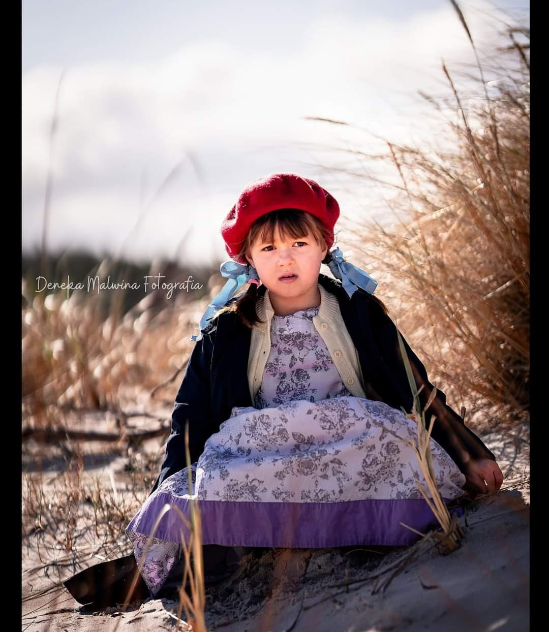 Portret dziewczynki w czerwonym berecie i kwiecistej sukience, siedzącej na piasku wśród wysokich traw, z delikatnym światłem podkreślającym jej twarz.