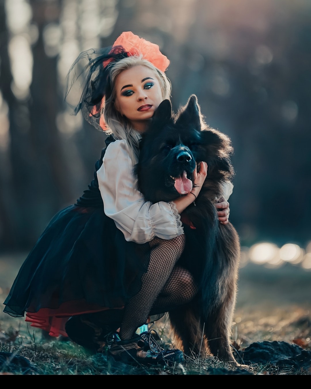 Kobieta w stylizowanej sukience i rajstopach w siatkę obejmuje dużego, czarnego psa na tle zamazanego, leśnego krajobrazu.