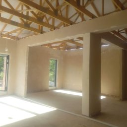 Tynki Maszynowe - Surowe wnętrze domu w trakcie tynkowania. Widoczne drewniane elementy konstrukcji dachu, ściany pokryte świeżym tynkiem, okna z widokiem na zieleń.