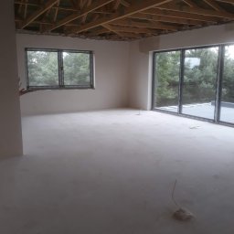 Tynki Maszynowe - Surowe wnętrze z oknami na las i drewnianą konstrukcją stropu. Wylewka podłogowa świeżo wykonana, widoczne wystające przewody elektryczne, ściany przygotowane do dalszych prac.