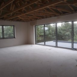 Tynki Maszynowe - Surowe wnętrze z dużymi oknami przesuwnymi i drewnianą konstrukcją sufitu. Podłoga przygotowana pod wykończenie, widoczne wystające przewody elektryczne.