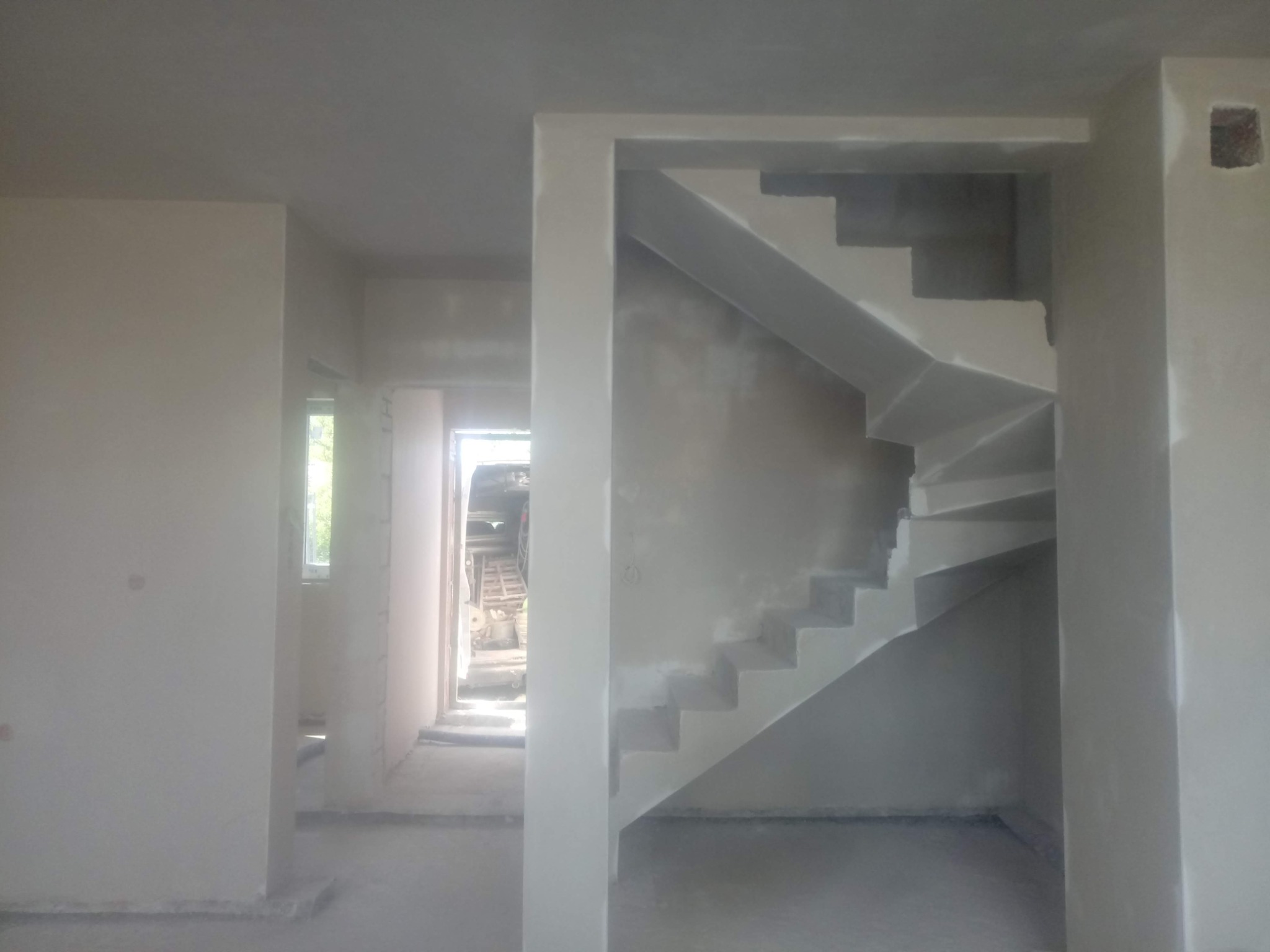 Wnętrze w trakcie wykończenia: surowe ściany, schody w stanie deweloperskim, widok na jasne pomieszczenie z oknem i sprzętem budowlanym w tle. Szary odcień ścian.