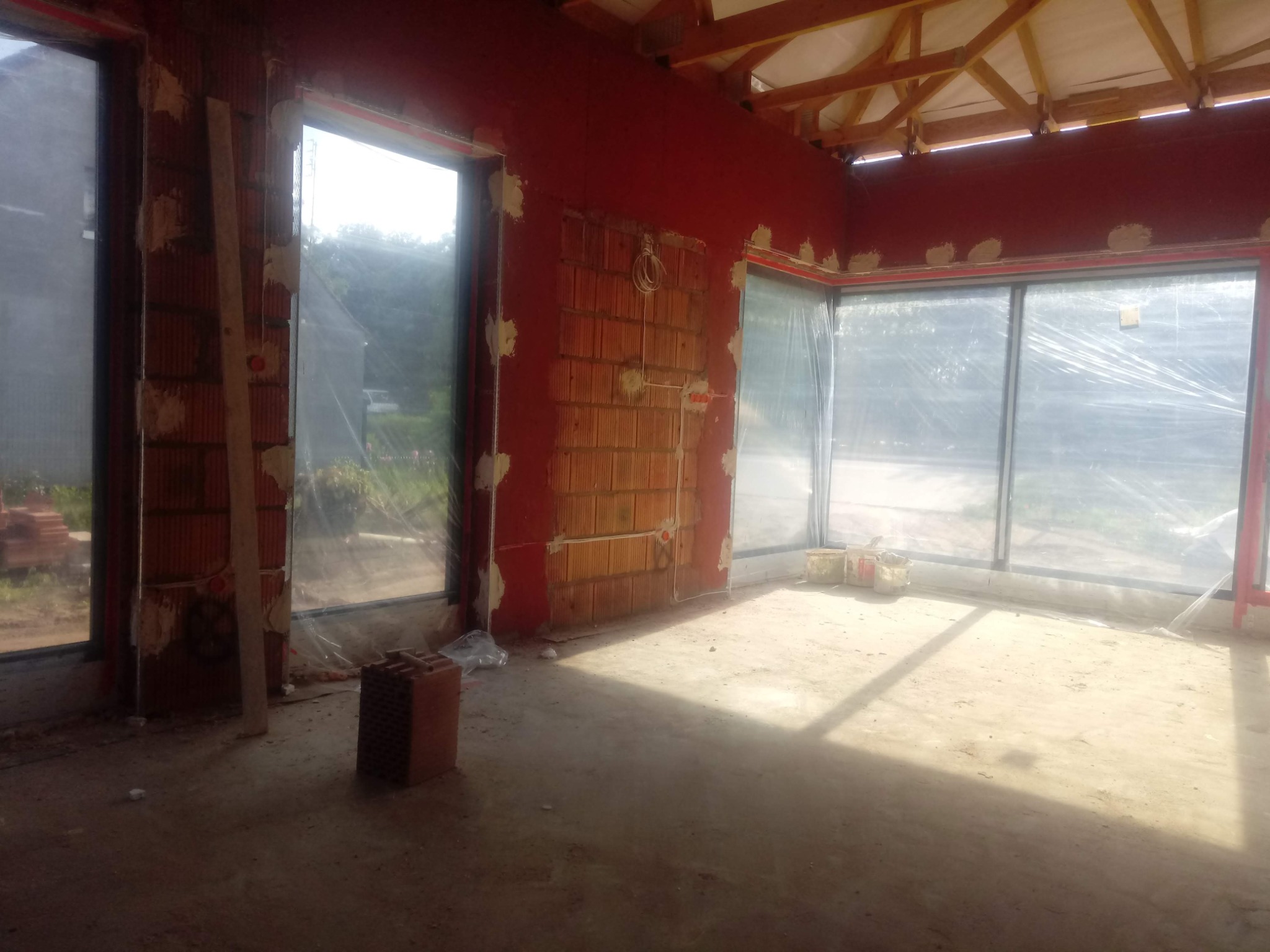 Surowy stan wnętrza w trakcie prac wykończeniowych: ściany z cegły, częściowo otynkowane, duże okna z folią ochronną, widoczne instalacje elektryczne, drewniana konstrukcja dachu.