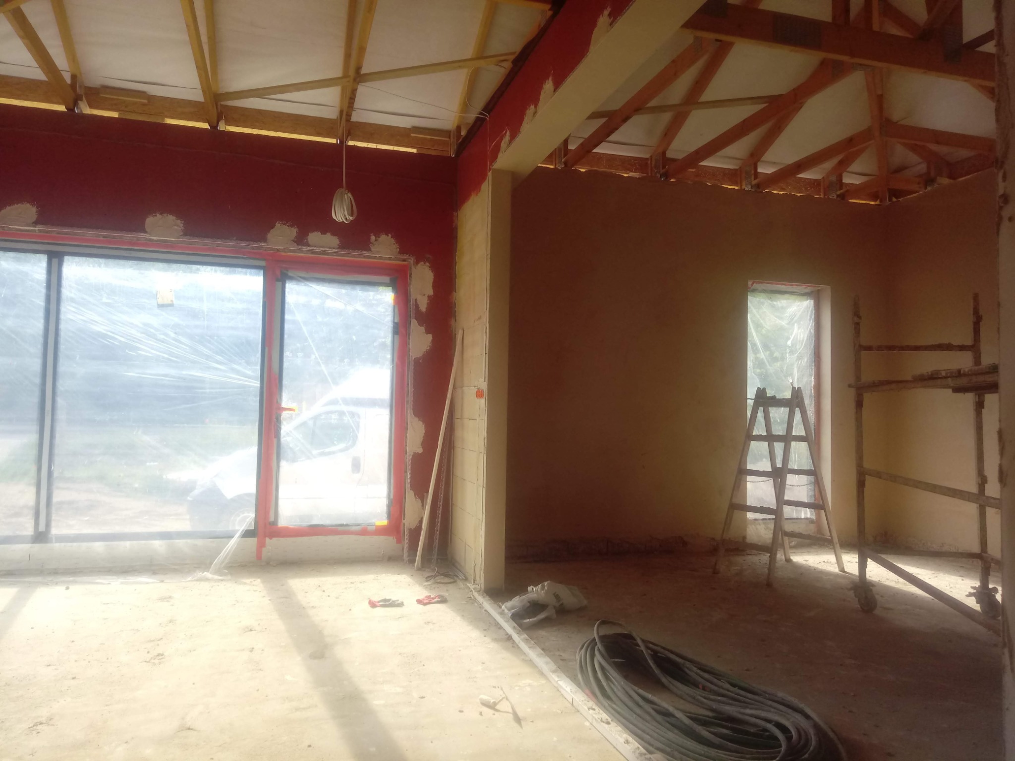 Wnętrze w trakcie tynkowania: czerwona ściana z widocznymi łatami tynku, okno zabezpieczone folią, drewniana konstrukcja sufitu, drabina i zwój kabli na podłodze.