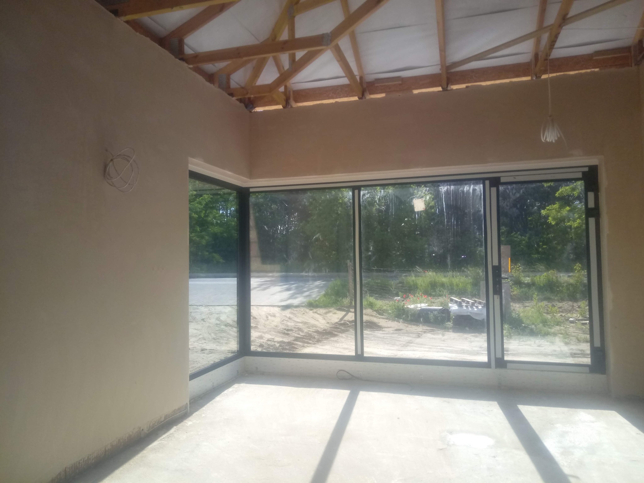 Wnętrze w stanie deweloperskim z dużym przeszkleniem. Ściany pokryte tynkiem, widoczne drewniane elementy konstrukcji dachu. Na ścianie zwisające przewody elektryczne.