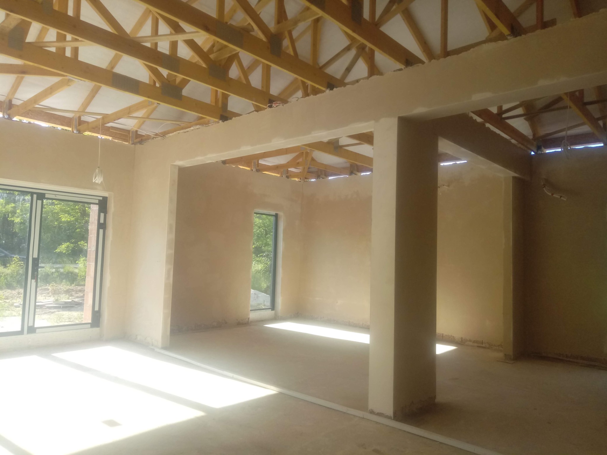 Surowe wnętrze domu w trakcie tynkowania. Widoczne drewniane elementy konstrukcji dachu, ściany pokryte świeżym tynkiem, okna z widokiem na zieleń.