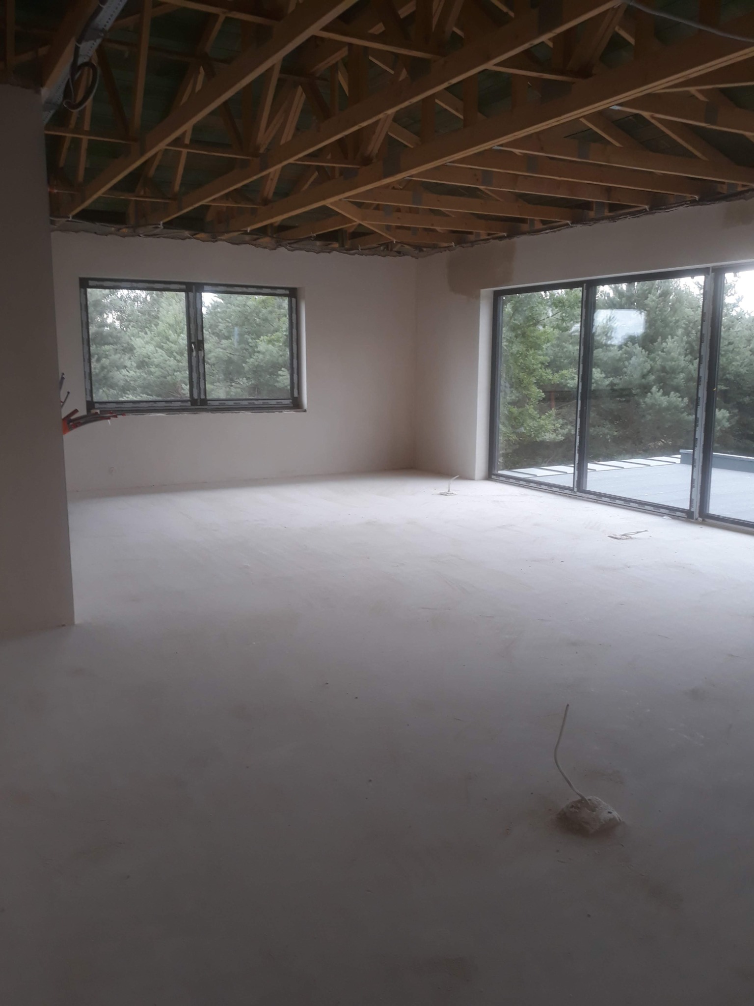 Surowe wnętrze z oknami na las i drewnianą konstrukcją stropu. Wylewka podłogowa świeżo wykonana, widoczne wystające przewody elektryczne, ściany przygotowane do dalszych prac.