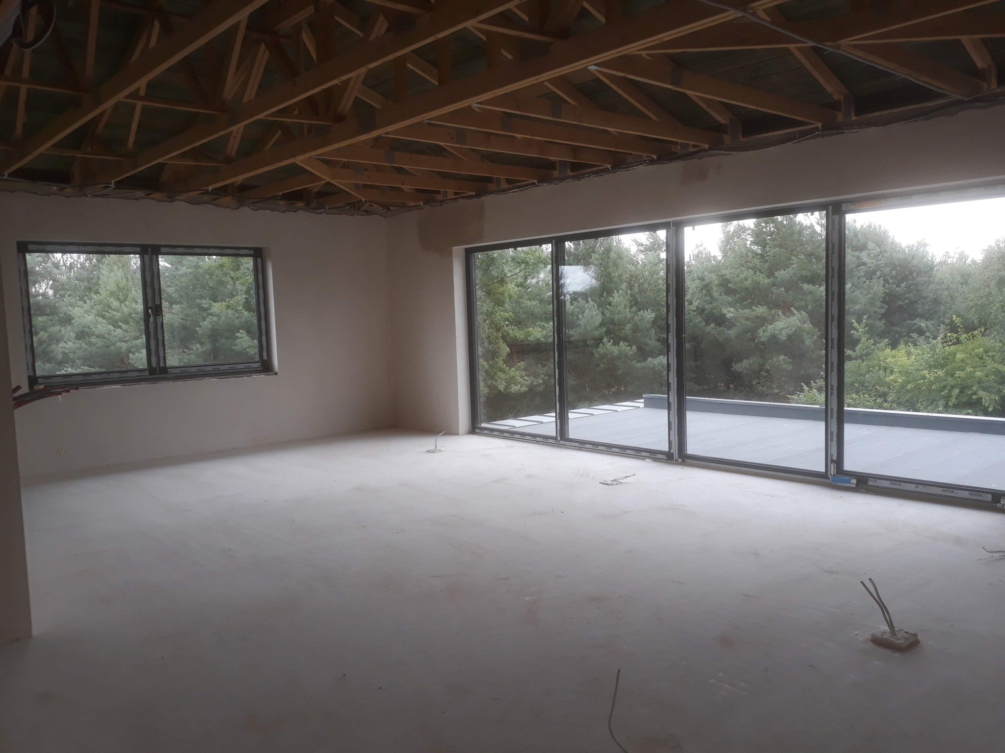 Surowe wnętrze z dużymi oknami przesuwnymi i drewnianą konstrukcją sufitu. Podłoga przygotowana pod wykończenie, widoczne wystające przewody elektryczne.