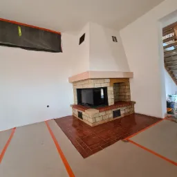 Wykończony kominek z cegły i kamienia w jasnym wnętrzu, podłoga częściowo zabezpieczona kartonem i taśmą malarską, widoczne ślady prac wykończeniowych.