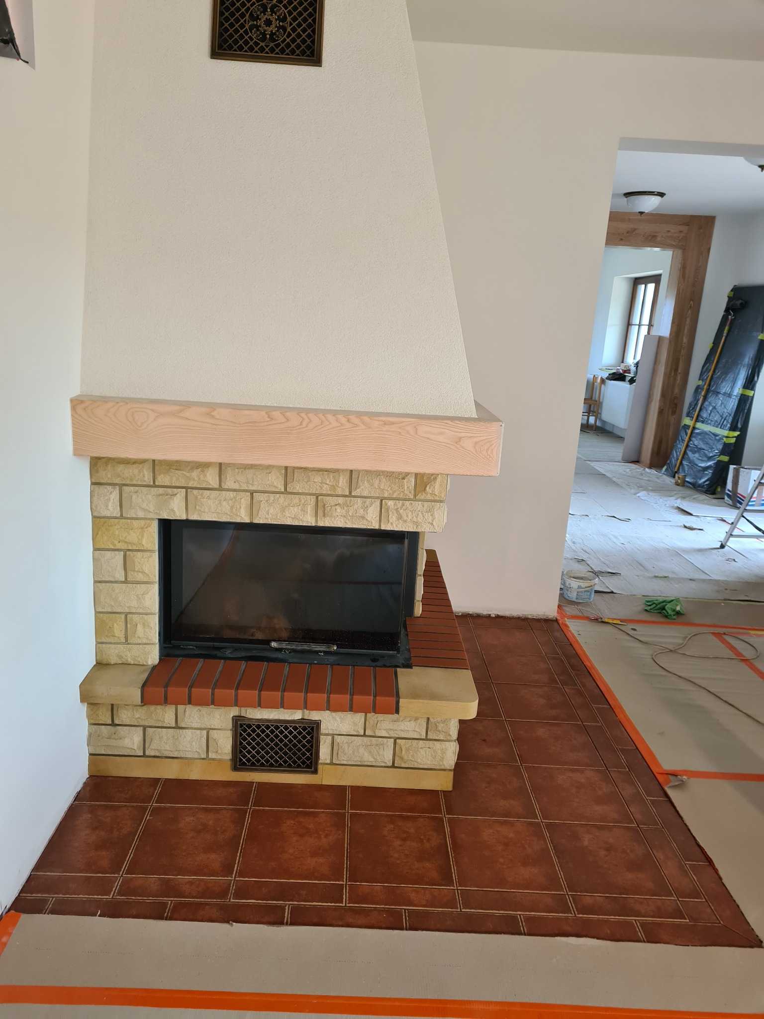 Wykończony kominek z jasnych cegieł i drewnianą półką, brązowa podłoga z płytek, widoczne zabezpieczenia podłogi na czas remontu, w tle otwarte przejście do innego pomieszczenia.