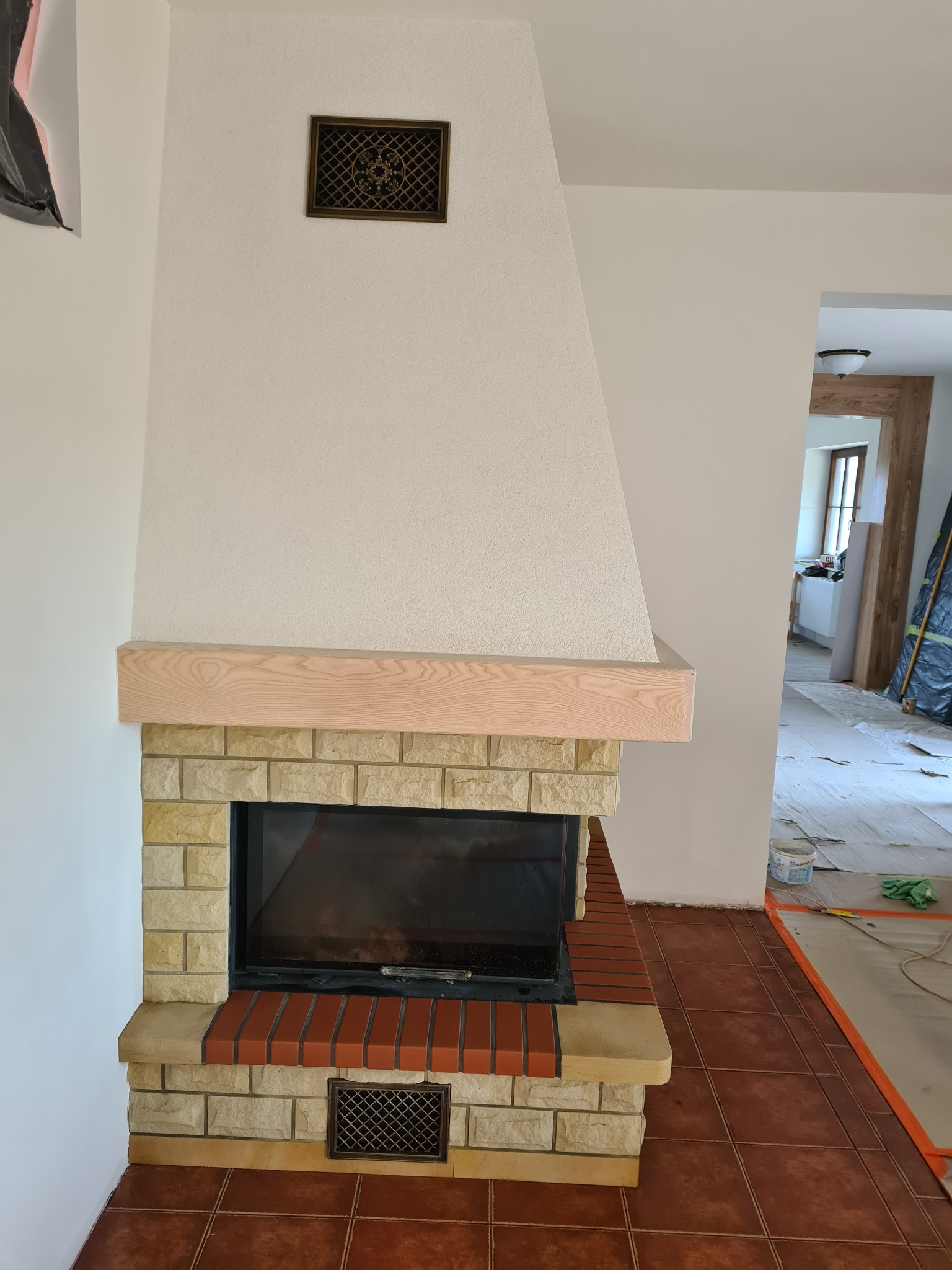 Nowoczesny kominek obudowany jasnym kamieniem i cegłą, z drewnianą półką, w trakcie prac wykończeniowych, widoczne zabezpieczenia podłogi i przejście do innego pomieszczenia.