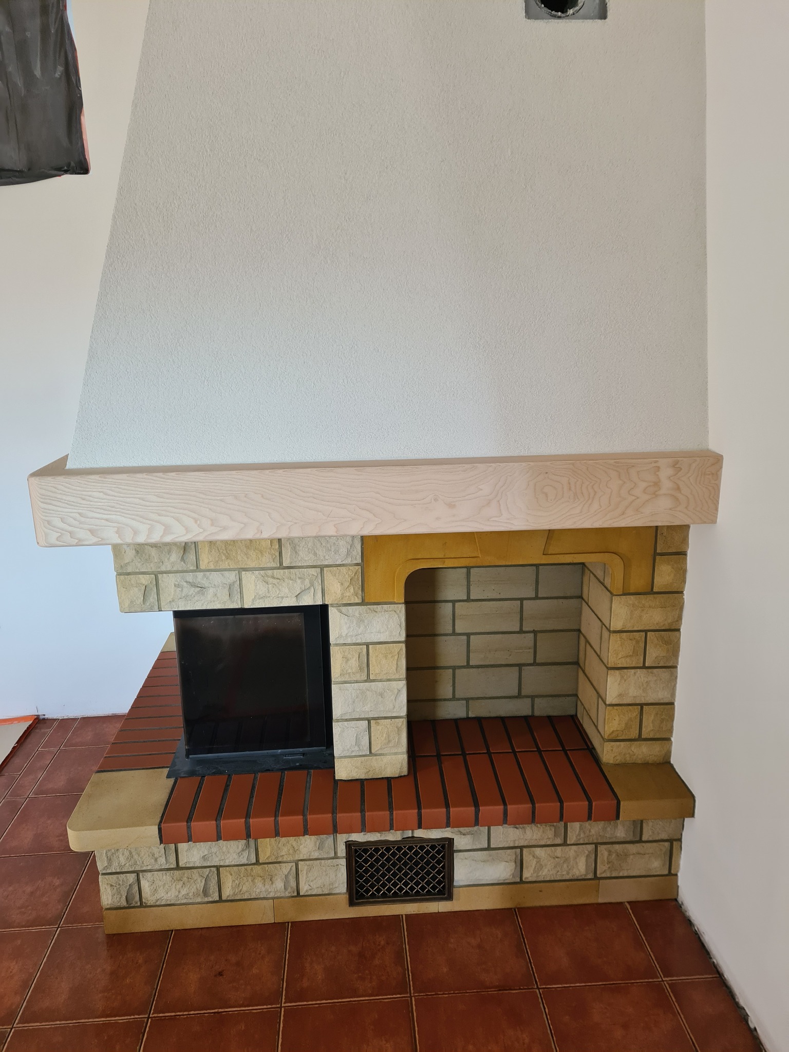 Wykończony kominek z obudową z kamienia i cegły, drewnianą półką i wkładem kominkowym, na podłodze terakota.