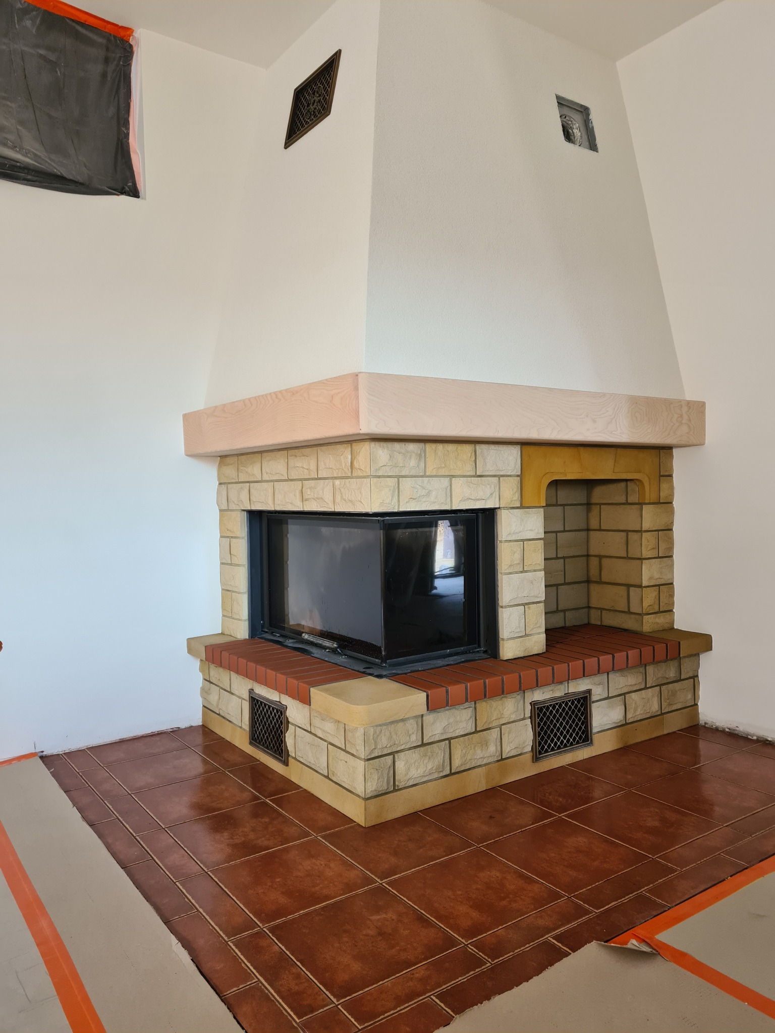 Nowoczesny kominek obłożony beżową cegłą i czerwoną cegłą klinkierową, z drewnianym blatem, widoczne kratki wentylacyjne, zabezpieczona podłoga z płytek ceramicznych.