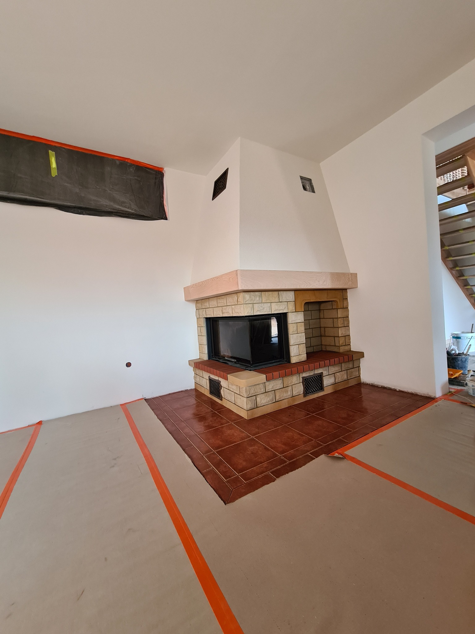 Wykończony kominek z cegły i kamienia w jasnym wnętrzu, podłoga częściowo zabezpieczona kartonem i taśmą malarską, widoczne ślady prac wykończeniowych.