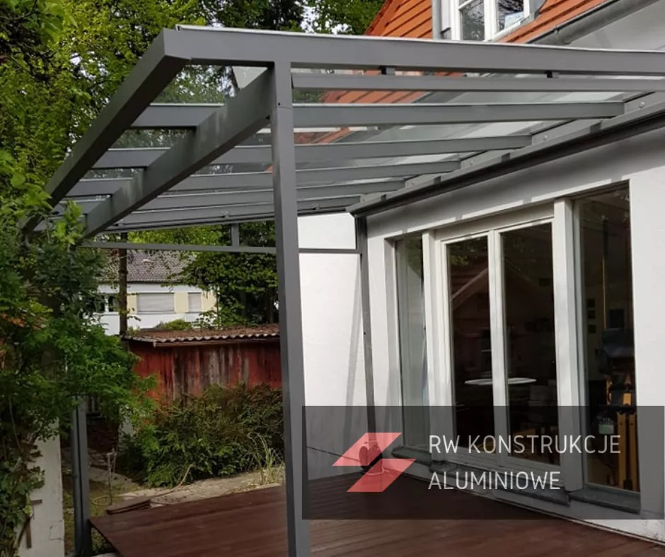 Szara konstrukcja zadaszenia tarasu z przezroczystym dachem, przylegająca do białego budynku z przesuwnymi drzwiami, widoczna z zewnątrz na tle zieleni ogrodu i drewnianego podestu.
