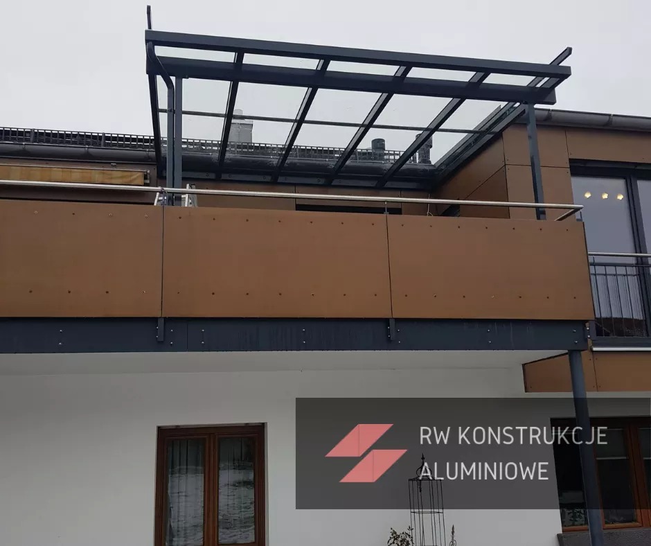 Zadaszenie balkonu z czarną konstrukcją stalową i przeszklonym dachem, widok z dołu na tle elewacji budynku z brązowymi panelami i białym murem.
