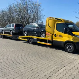 Żółta laweta marki Iveco z trzema samochodami (biały, szary SUV, czarny kombi) na platformie, zaparkowana na brukowanym placu w słoneczny dzień. Widoczne logo firmy 'AUTODANIEL' i napis 'MIASTKO'...