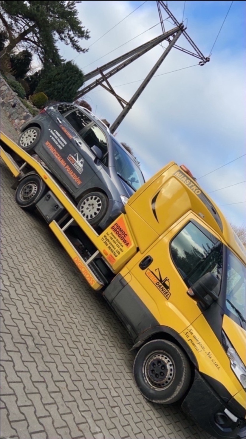 Żółta ciężarówka pomocy drogowej z szarym samochodem osobowym na platformie, widoczne logo firmy 'Auto Daniel' i hasło 'Na miejsce. Na czas. Na pewno!'