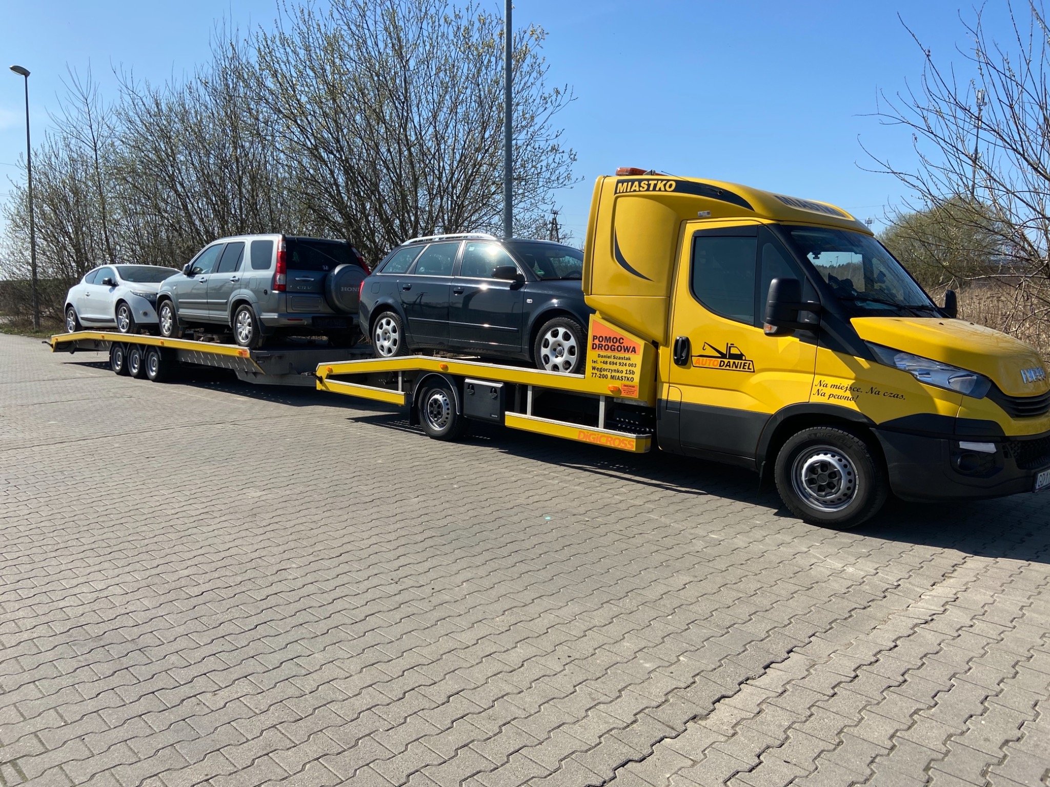 Żółta laweta marki Iveco z trzema samochodami (biały, szary SUV, czarny kombi) na platformie, zaparkowana na brukowanym placu w słoneczny dzień. Widoczne logo firmy 'AUTODANIEL' i napis 'MIASTKO'...
