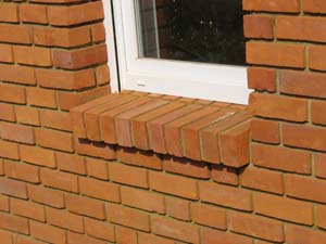 Fragment elewacji z czerwonej cegły klinkierowej z białym oknem i ozdobnym ceglanym parapetem, widoczne spoiny między cegłami.