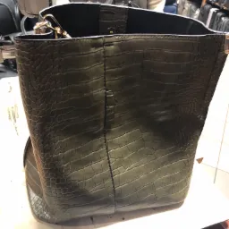 Duża, otwarta torba damska w kolorze khaki, imitująca skórę krokodyla, z metalowymi elementami, stojąca na stole w warsztacie, obok maszyny do szycia.