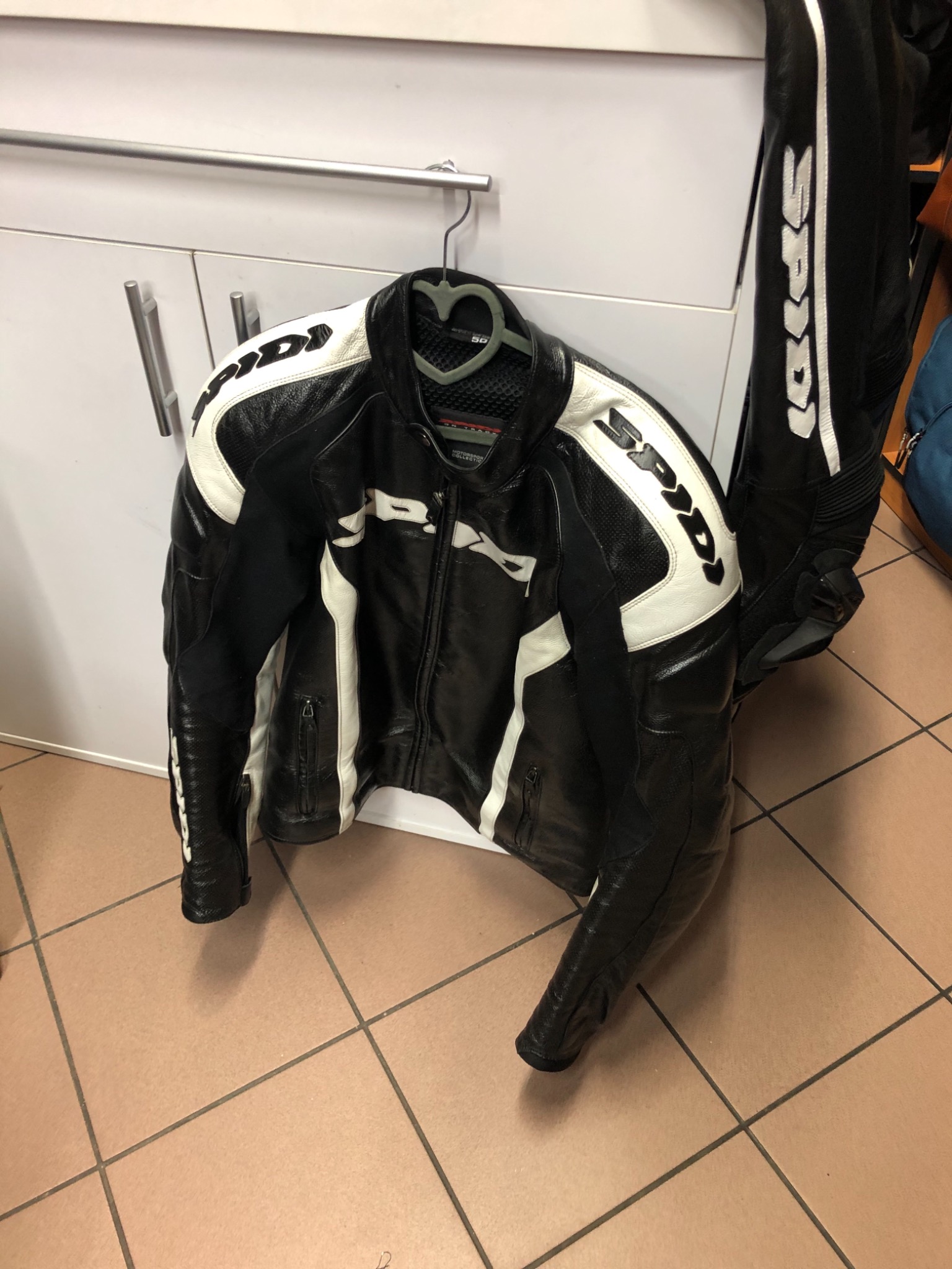 Czarno-biała skórzana kurtka motocyklowa marki SPIDI zawieszona na wieszaku, obok spodnie motocyklowe, widoczne na tle białych szafek i płytek podłogowych.