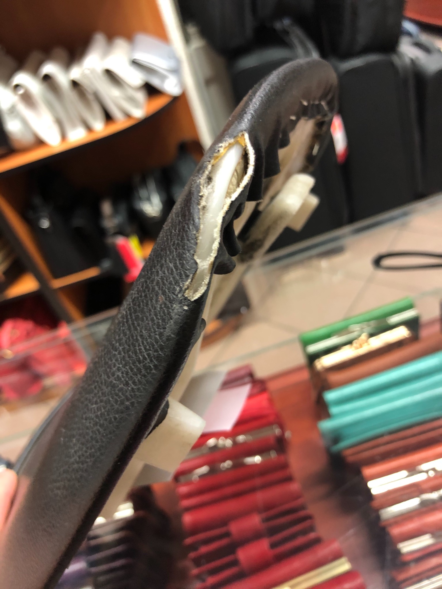 Uszkodzony, czarny pasek skórzany z widocznym białym wypełnieniem, leżący na szklanej powierzchni, w tle inne wyroby skórzane.