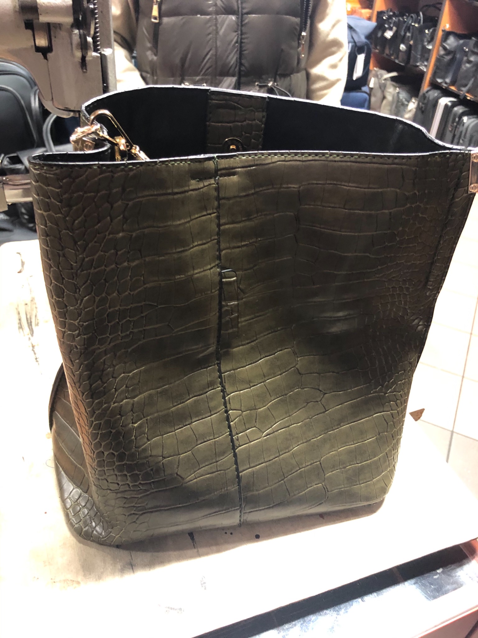 Duża, otwarta torba damska w kolorze khaki, imitująca skórę krokodyla, z metalowymi elementami, stojąca na stole w warsztacie, obok maszyny do szycia.