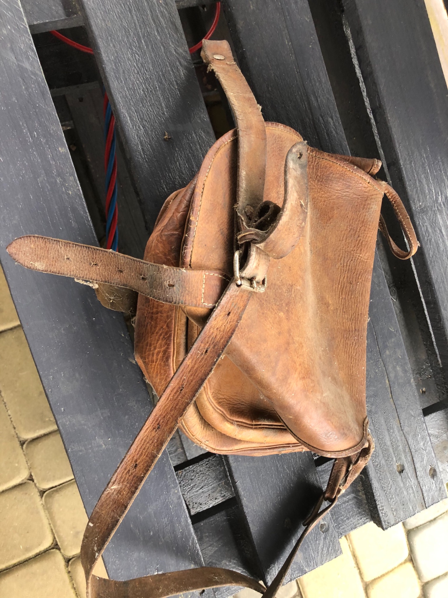 Stara, skórzana torba z długim paskiem leży na czarnych, drewnianych deskach. Widoczne ślady użytkowania i przetarcia na skórze.