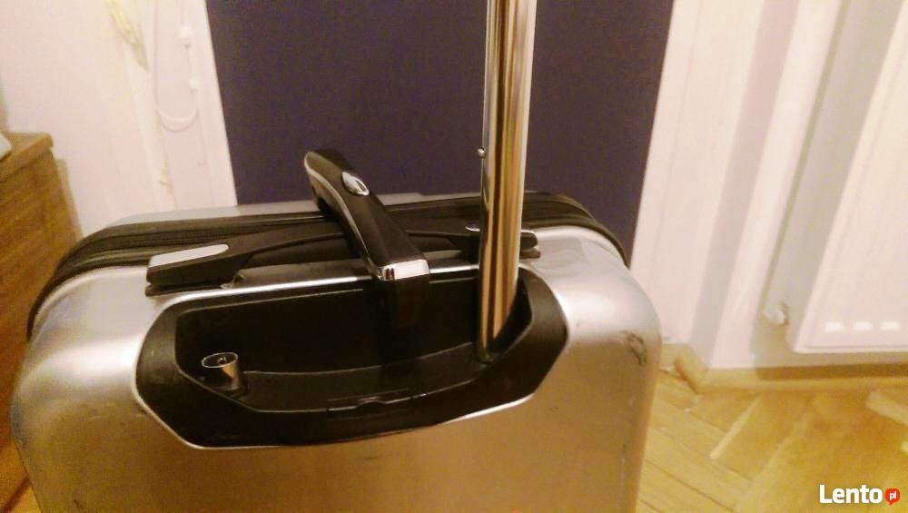 Błyszcząca, srebrna walizka na kółkach z wysuwaną rączką, widoczna na tle ściany w odcieniach szarości i bieli.