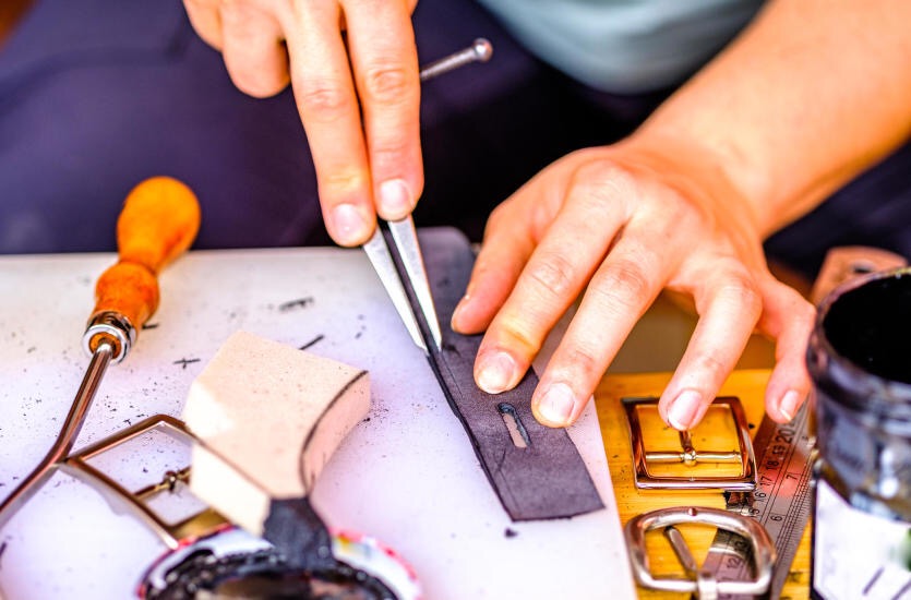 Przygotowanie skórzanego paska do sprzączki: Ręczne wycinanie otworu w ciemnoszarym pasku skórzanym za pomocą metalowych szczypiec na białym podłożu, widoczne narzędzia kaletnicze i metalowe...